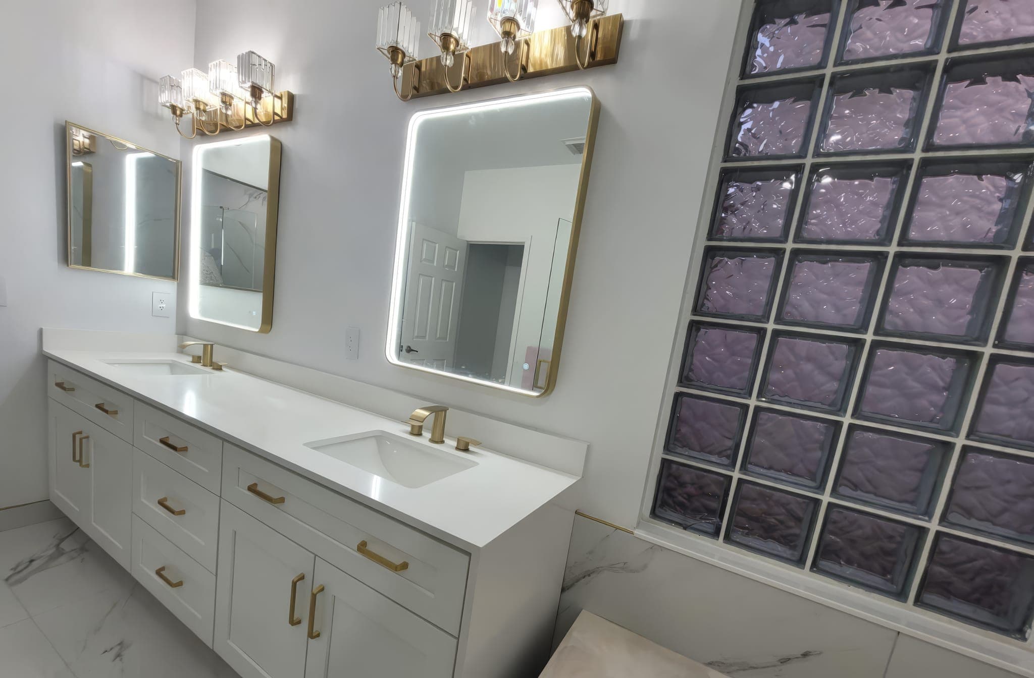 Double vanity with gold faucets, lit mirrors and glass block window