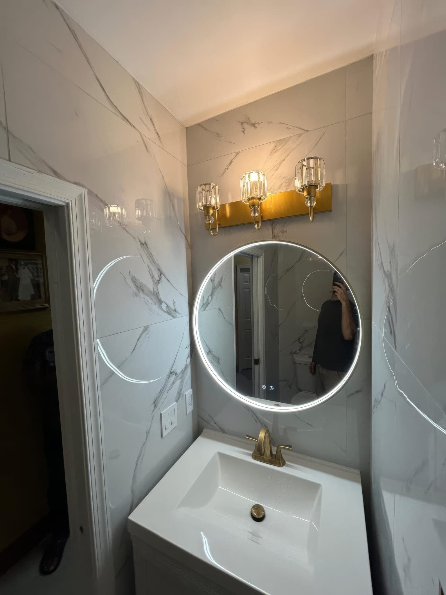 Round lit mirror and gold sconce over a white vanity