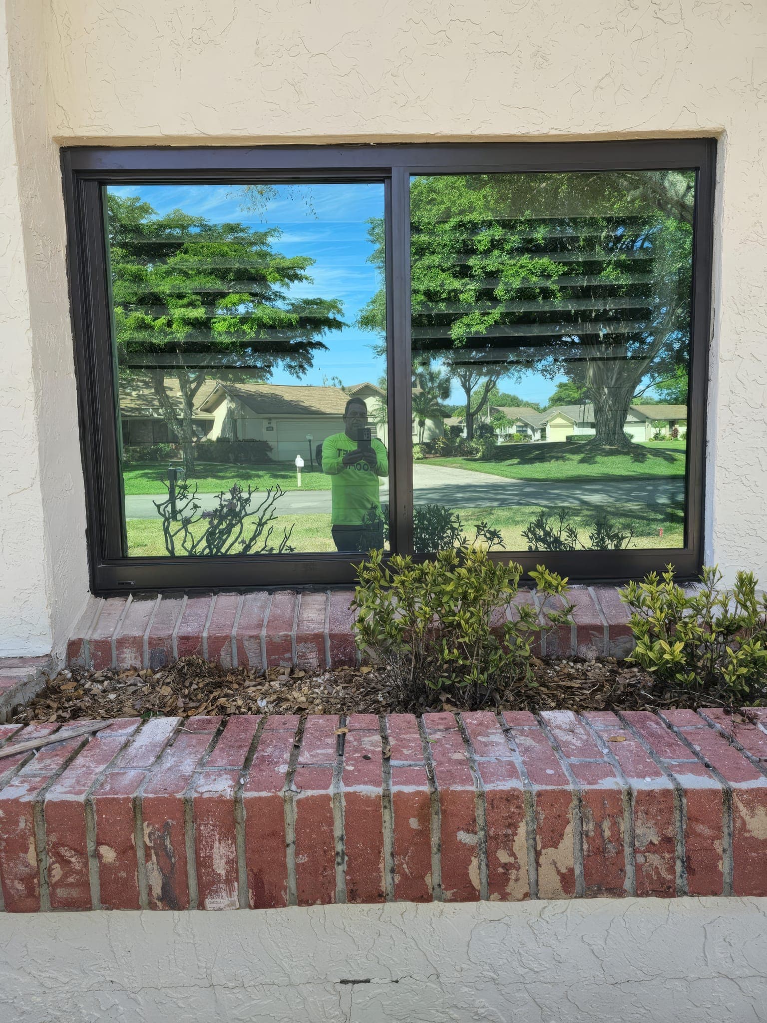 New bronze impact slider window above the brick planter, reflecting the yard